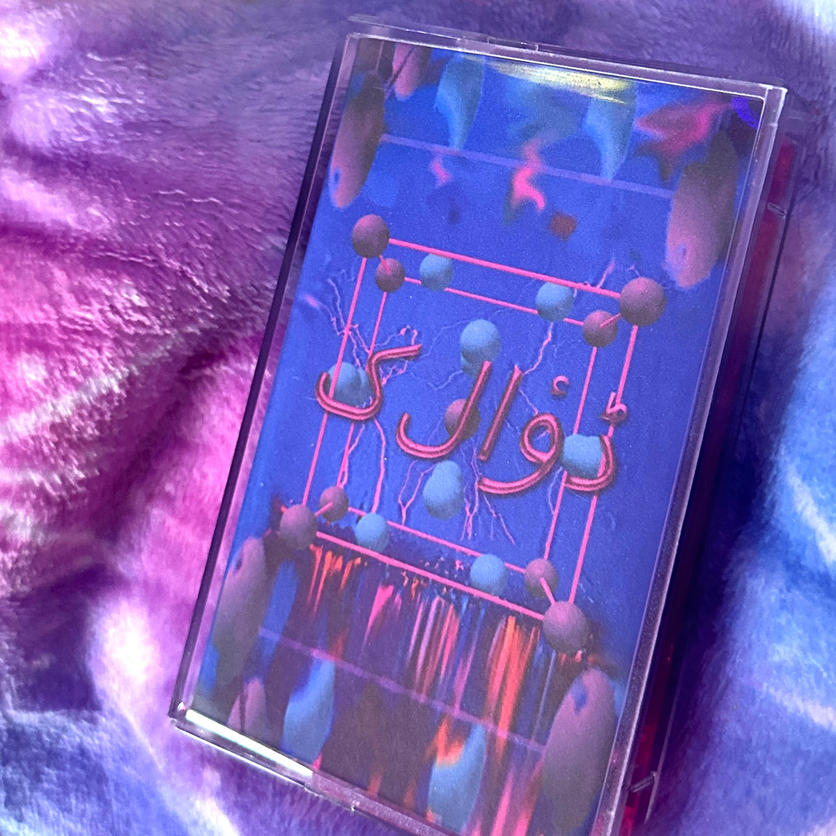 A clear plastic cassette-style case containing a blue album or book cover with Arabic text and abstract neon-like graphics, placed on a purple and blue textured fabric background.