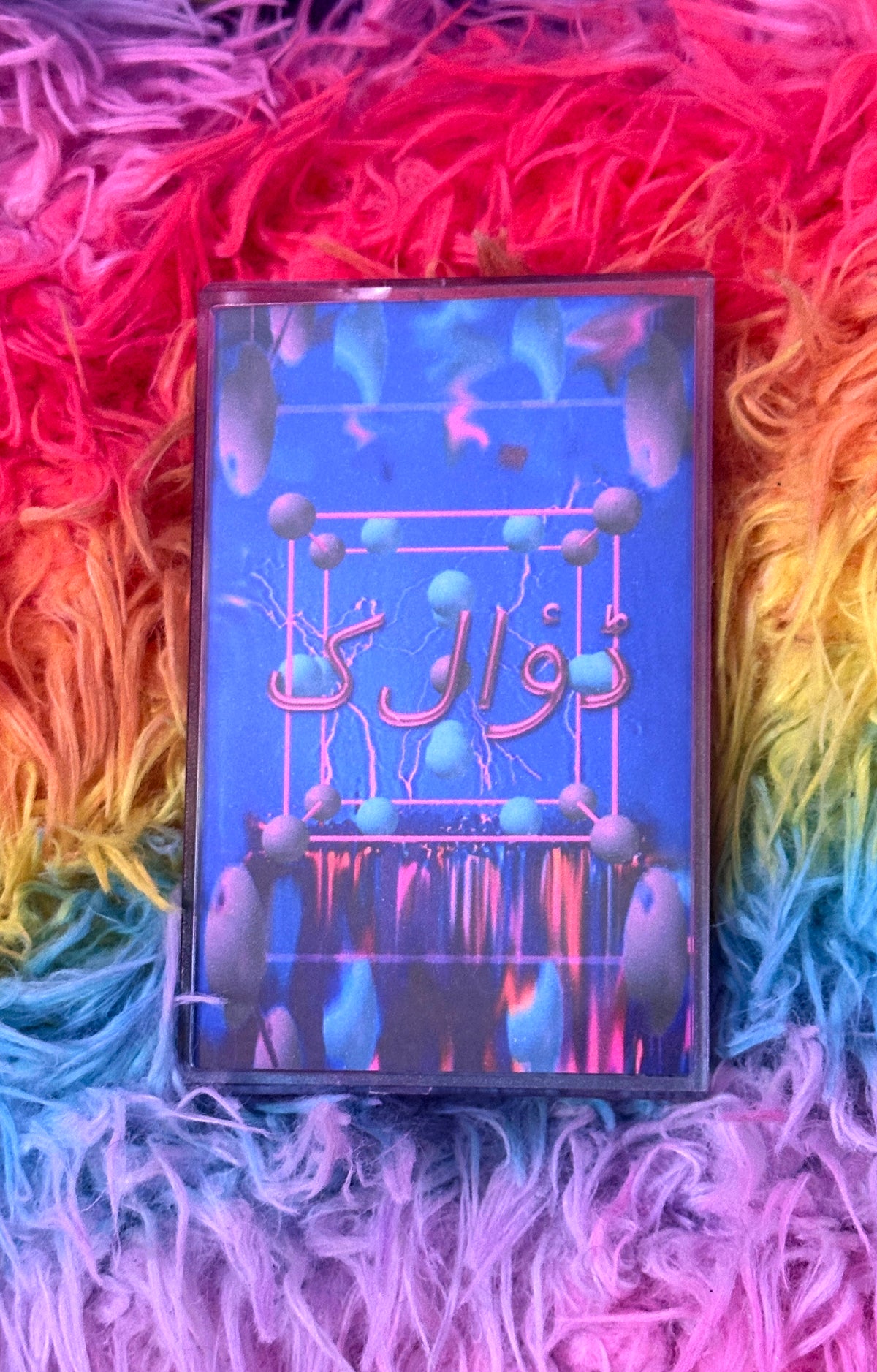 A cassette-style music album cover or book-style package with Arabic text, placed on a bright rainbow-colored shag rug. The cover is predominantly blue with pink and purple abstract graphics and text.